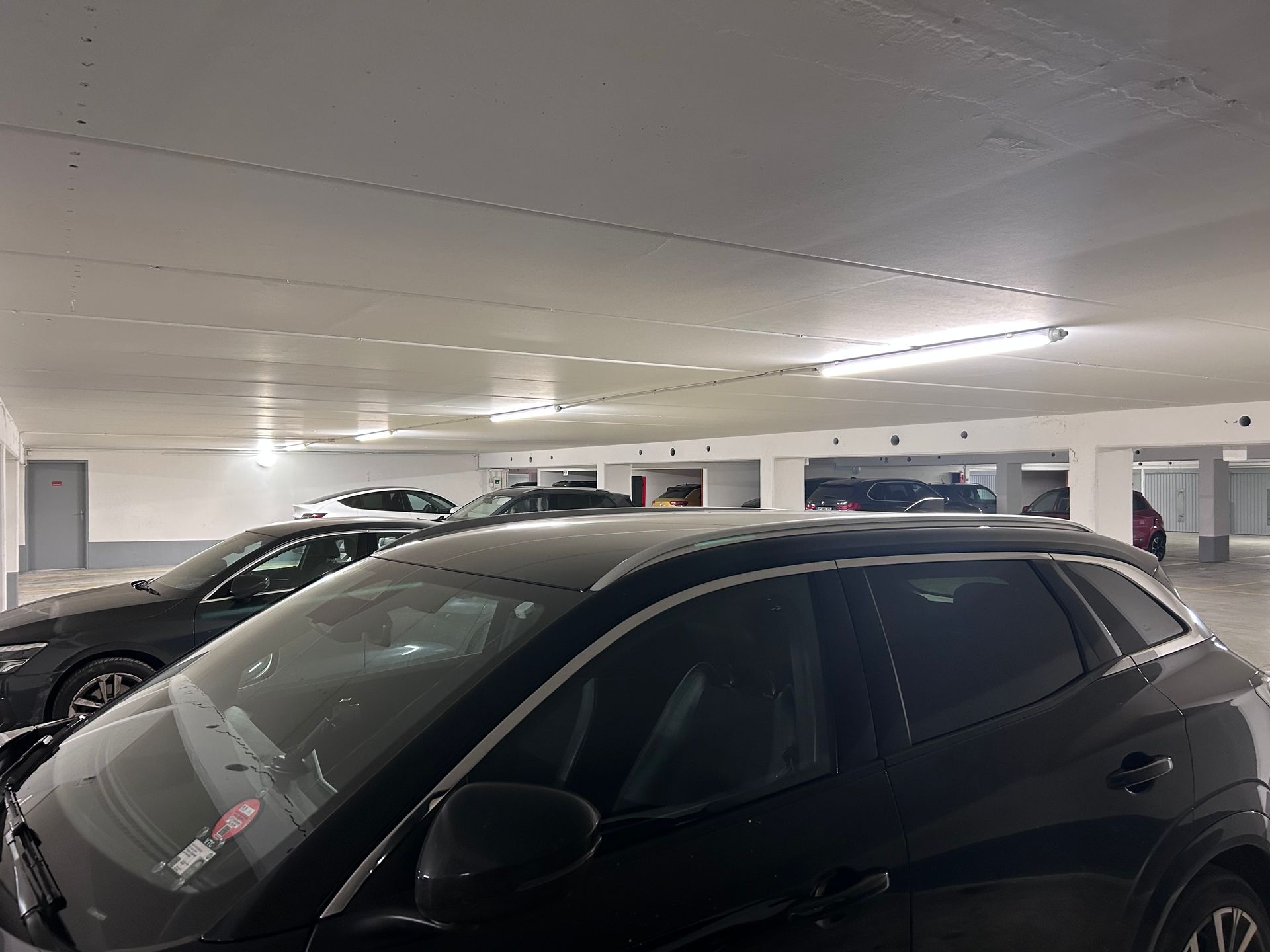 Voitures noires garées dans un parking intérieur éclairé par des plafonniers blancs.