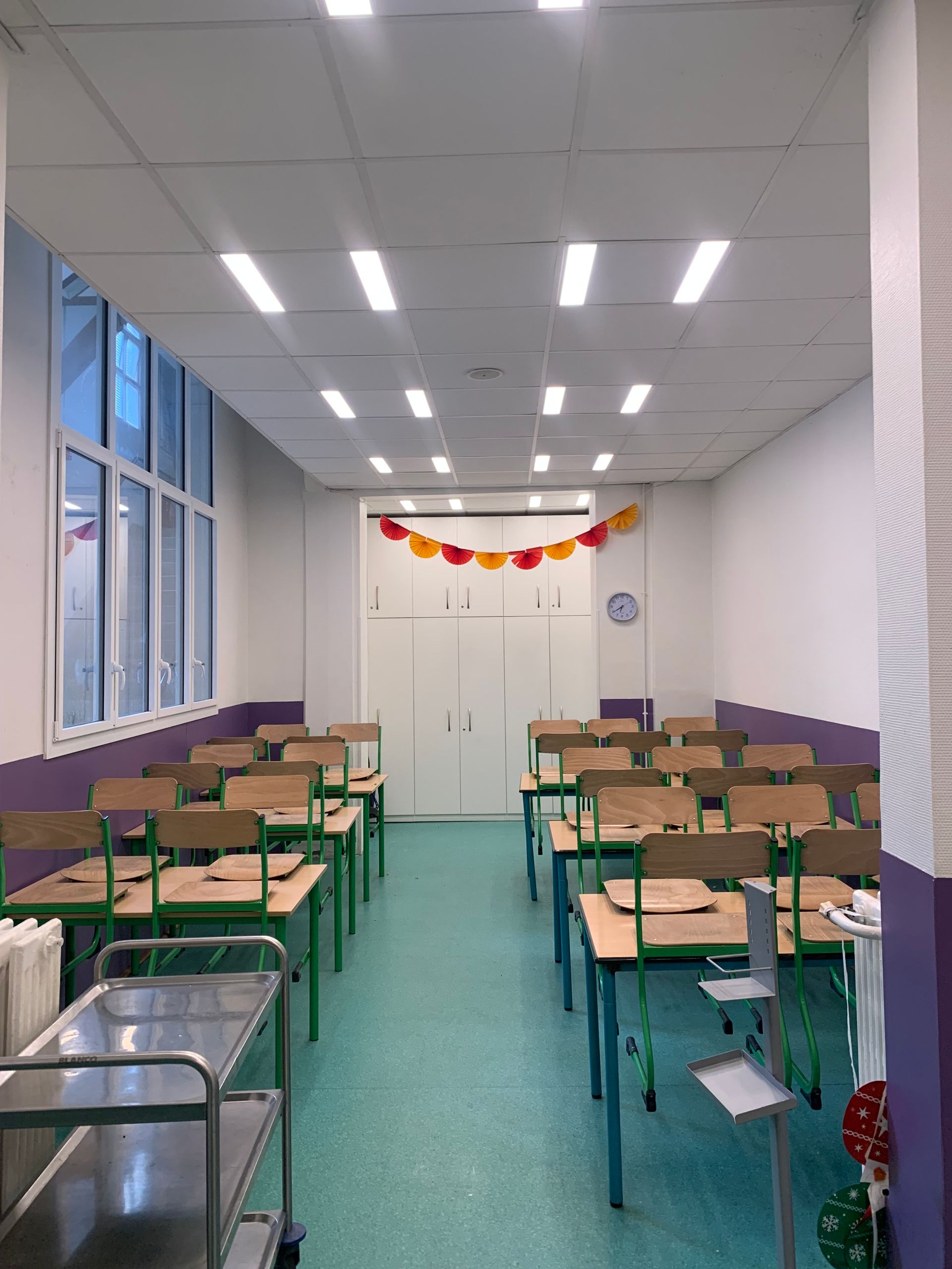 Salle de réfectoire vide avec des rangées de pupitres, un sol turquoise et des décorations colorées au-dessus des portes d'entrée.