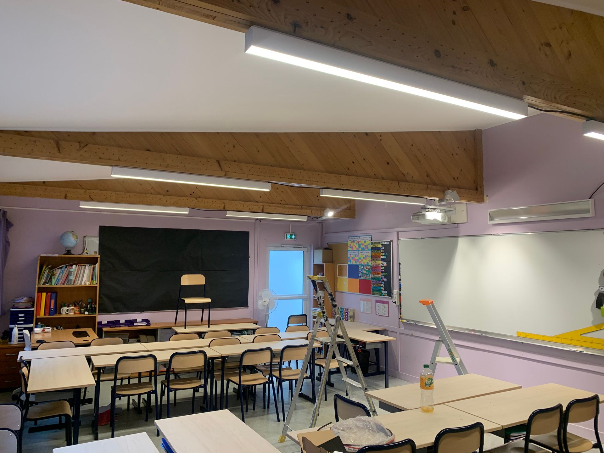 Salle de classe avec des rangées de pupitres, un tableau noir et une échelle sous des poutres apparentes au plafond.