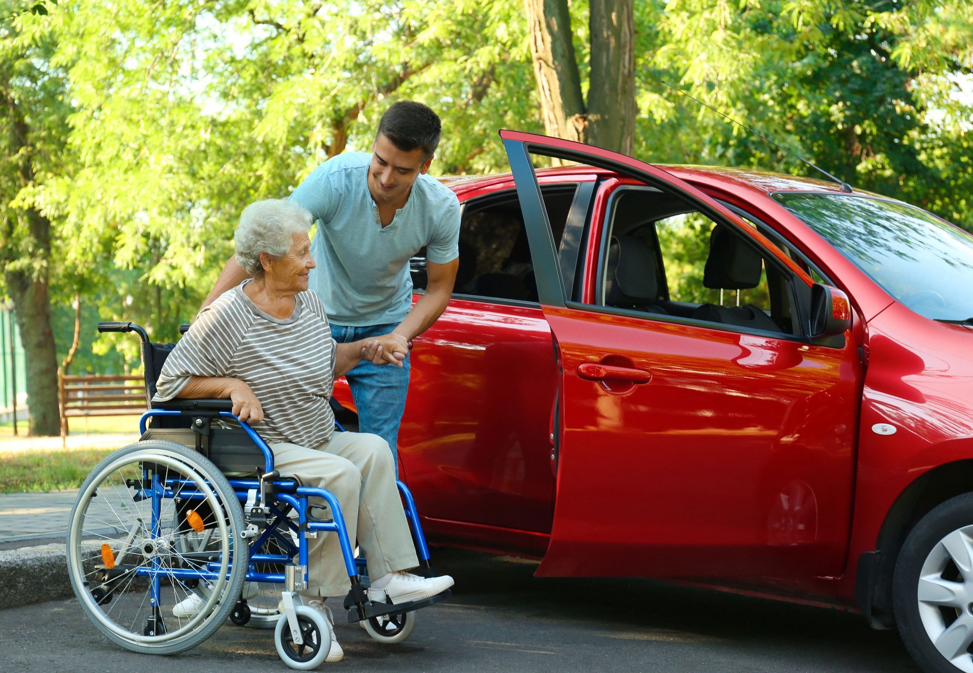 Un homme aide une femme âgée en fauteuil roulant à monter dans une voiture rouge. Extérieur, journée ensoleillée.