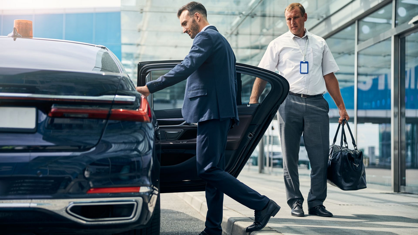 Un homme d'affaires sort d'une berline sombre ; un conducteur tient la porte et un sac à l'extérieur d'un bâtiment.