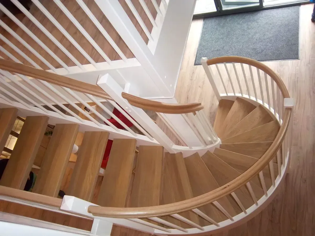 Draufsicht auf eine geschwungene Holztreppe mit weißen Geländern und Handläufen, die zu einem hellen Boden führt.