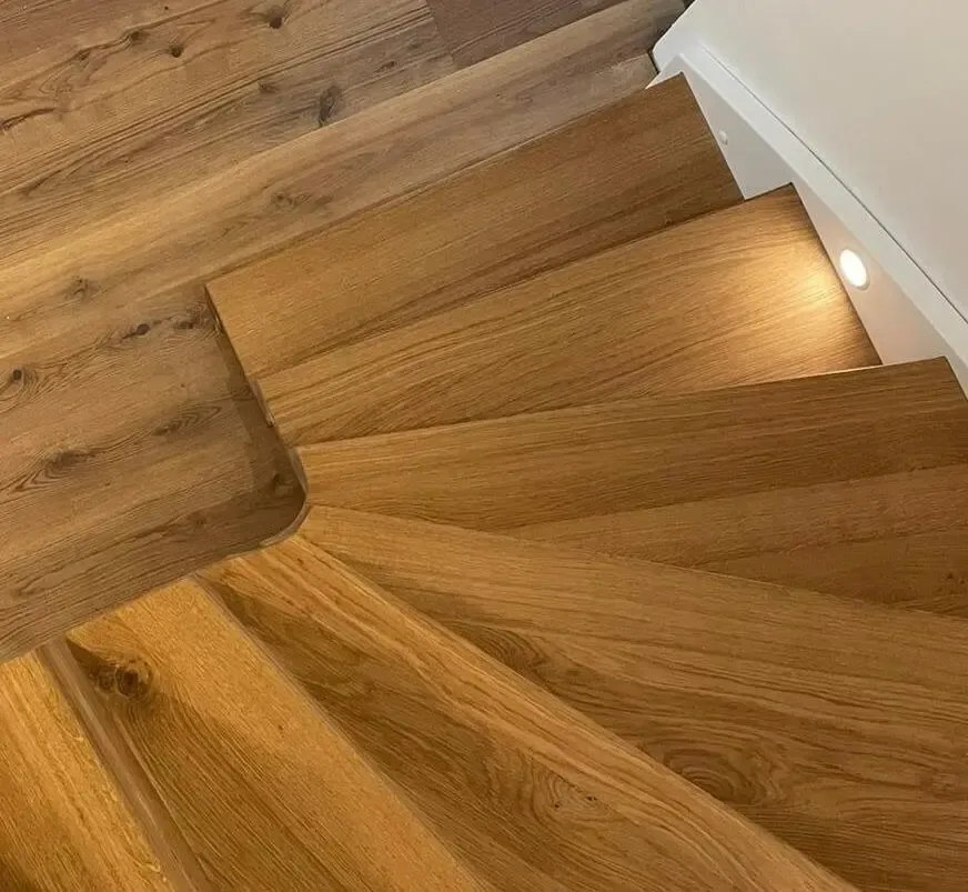 Eine hölzerne Treppe, die sich neben einer weißen Wand mit einer kleinen, runden Einbauleuchte nach unten windet.