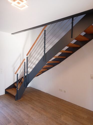 Moderne Treppe mit Holzstufen und dunklem Metallrahmen, Kabelgeländer und hellem Holzhandlauf.