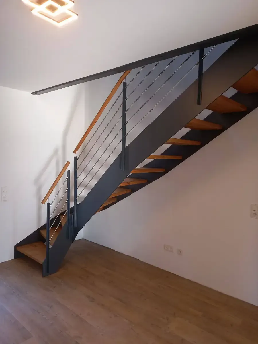 Moderne Treppe mit dunklen Metallwangen, Holzstufen und einem Kabelgeländer vor einer weißen Wand.