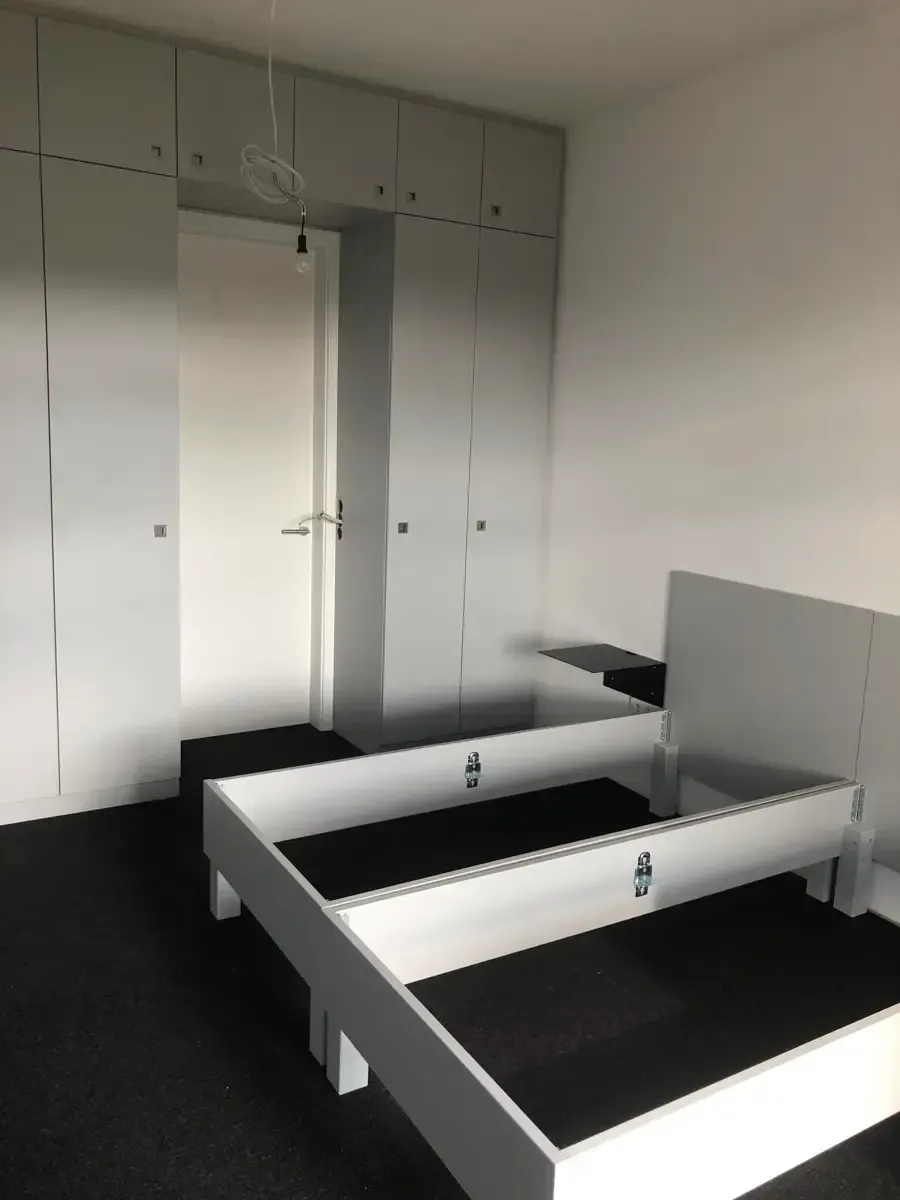 Ein minimalistisches Schlafzimmer mit weißem Bettgestell auf dunklem Teppich und hellgrauen, deckenhohen Einbauschränken.