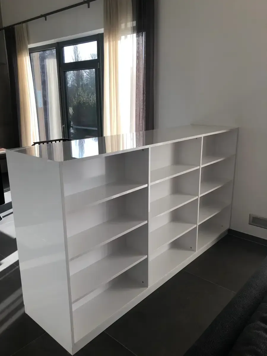 In einem Zimmer mit Fenster und Vorhängen im Hintergrund steht ein großes, weißes Bücherregal mit mehreren Regalböden.