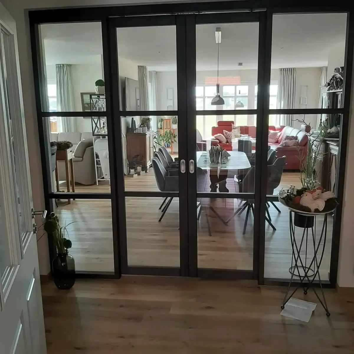 Schwarz gerahmte französische Glastüren führen in ein helles Wohn- und Esszimmer mit Holzböden und roten Möbeln.