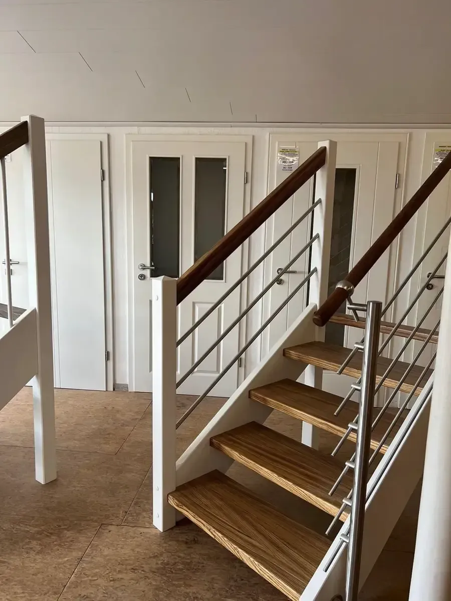 Eine Holztreppe mit weißem Geländer und Metallbalustern führt in einen Flur mit drei weißen Türen nach oben.