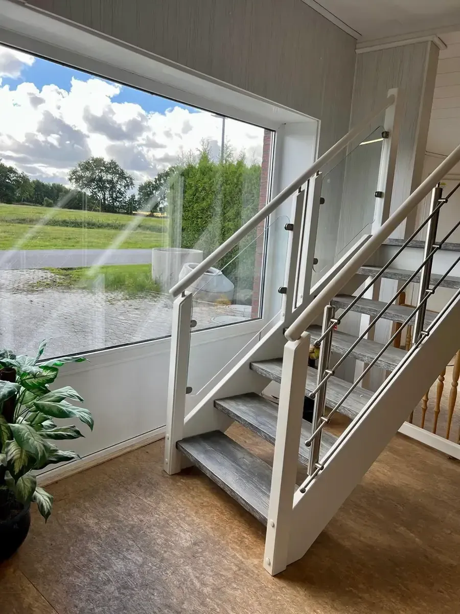Eine Treppe mit weißen Rahmen und Holzstufen, Glaspaneelen und Metallgeländern, neben einem großen Fenster.