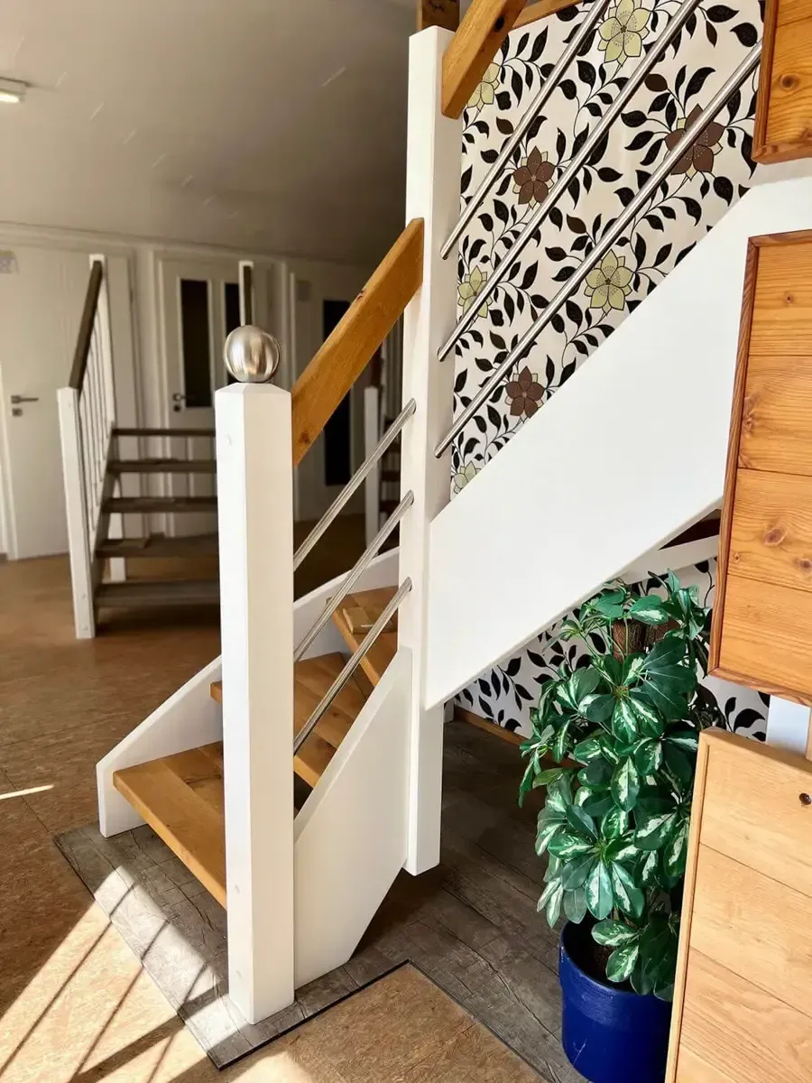 Eine Holztreppe mit weißem Rahmen und Metallgeländer, vor einer gemusterten, tapezierten Wand neben einer Topfpflanze.