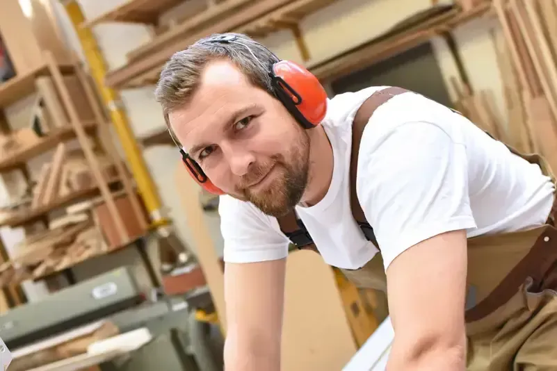 Ein Schreiner, der Gehörschutz und Arbeitsoverall trägt, schaut in die Kamera, während er in einer Holzwerkstatt arbeitet.