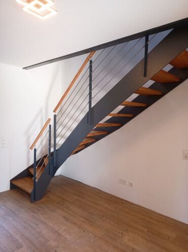 Moderne Treppe mit Holzstufen, dunklem Metallrahmen und Drahtgeländer in einem minimalistischen Raum mit Holzboden.