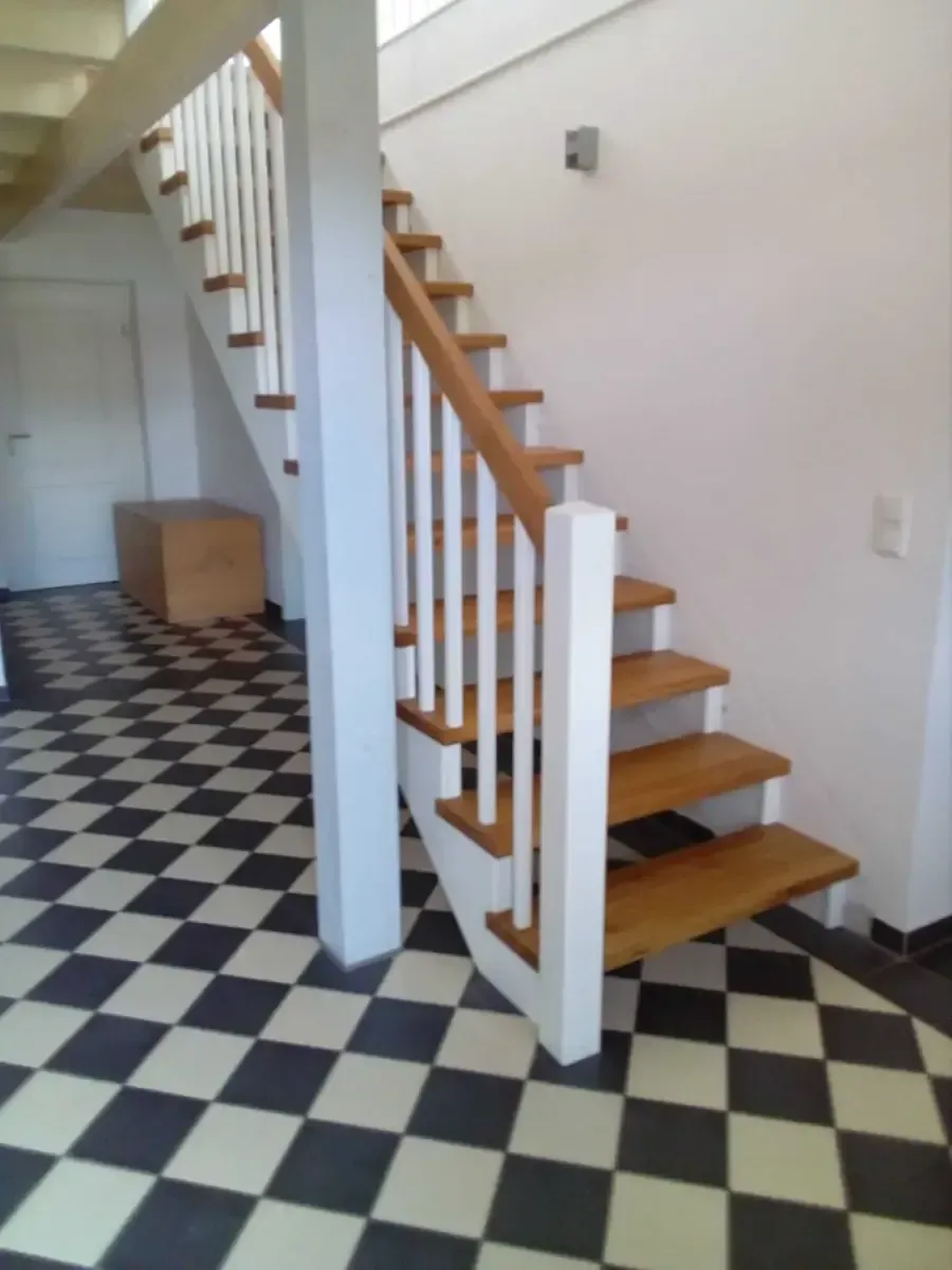 Treppe mit Holzstufen und weißem Geländer über einem schwarz-weiß karierten Fliesenboden.