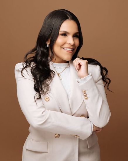 Una mujer con una chaqueta blanca y un suéter blanco sonríe con los brazos cruzados.