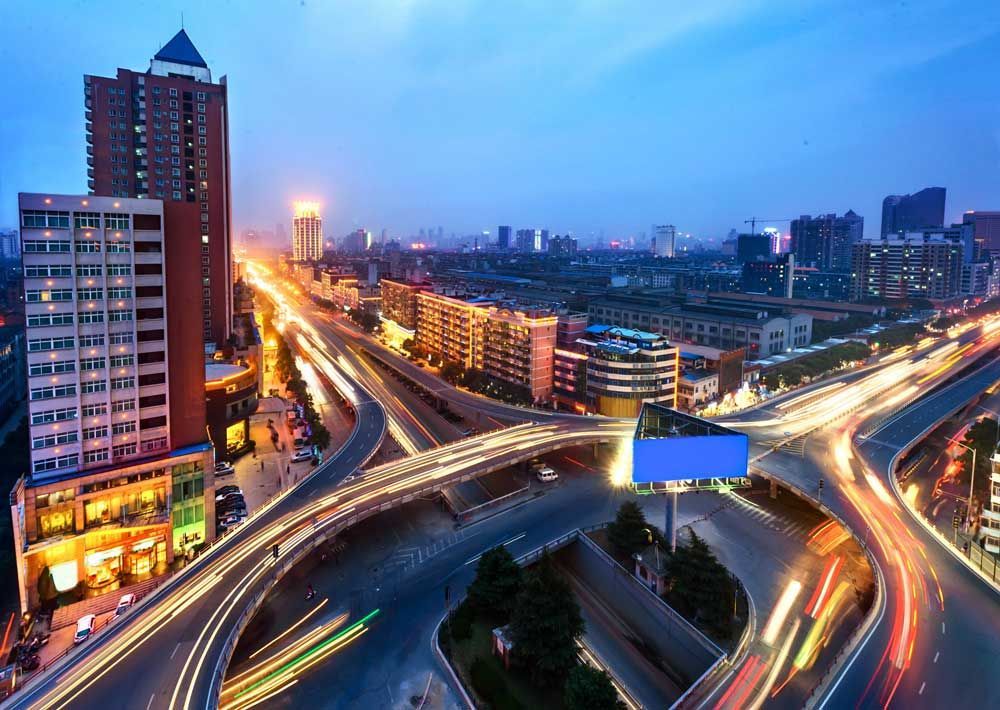 Intercambio de autopista elevada al anochecer; edificios de la ciudad con luces, rayas borrosas de tráfico y cielo azul.