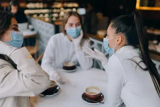 Tres mujeres con mascarillas y guantes están sentadas en una mesa con tazas de café.