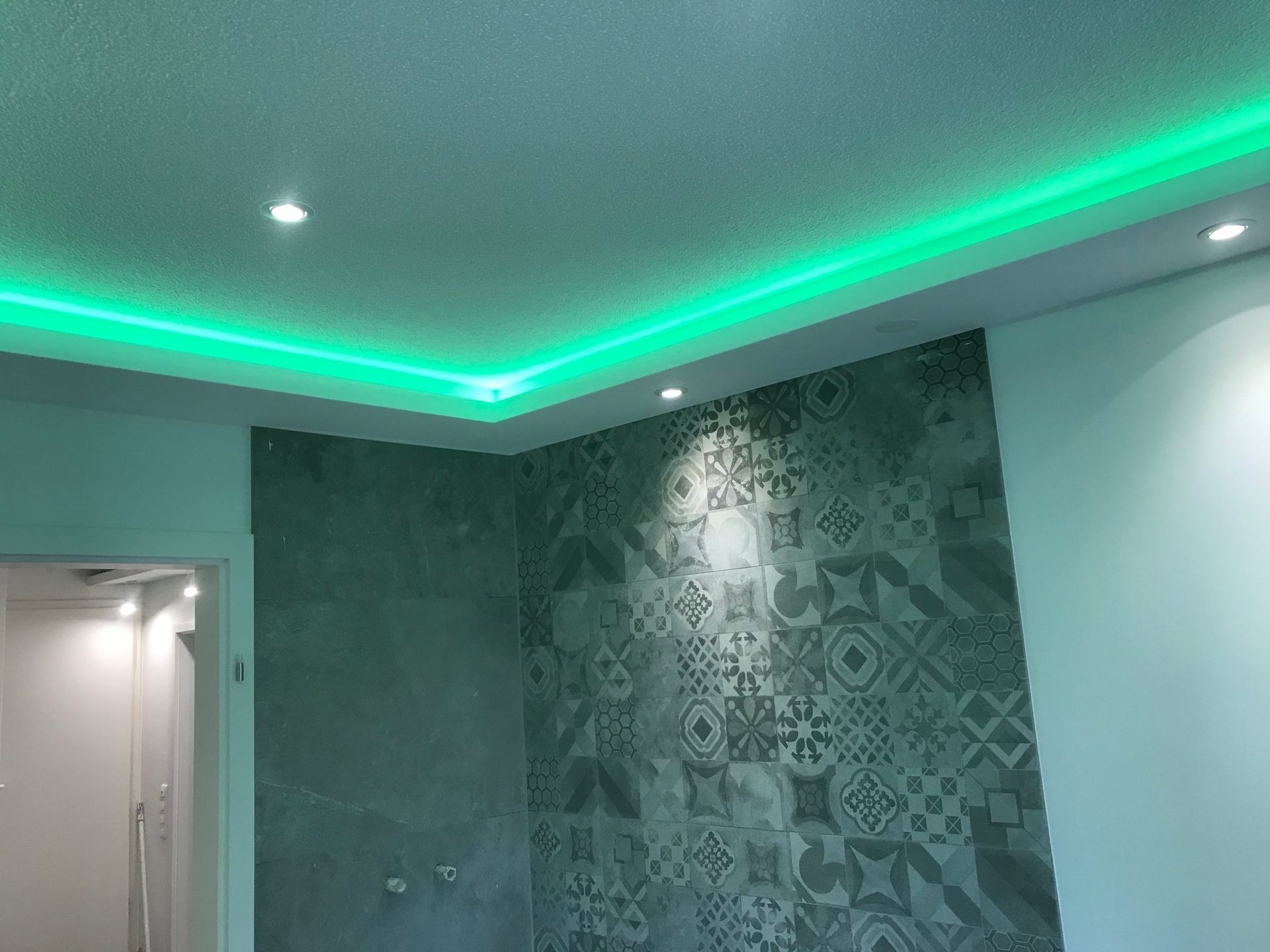 Grüne LED-Streifenbeleuchtung einer weißen Decke mit eingelassenen Strahlern in einem Raum mit Fliesen und einem Türrahmen.