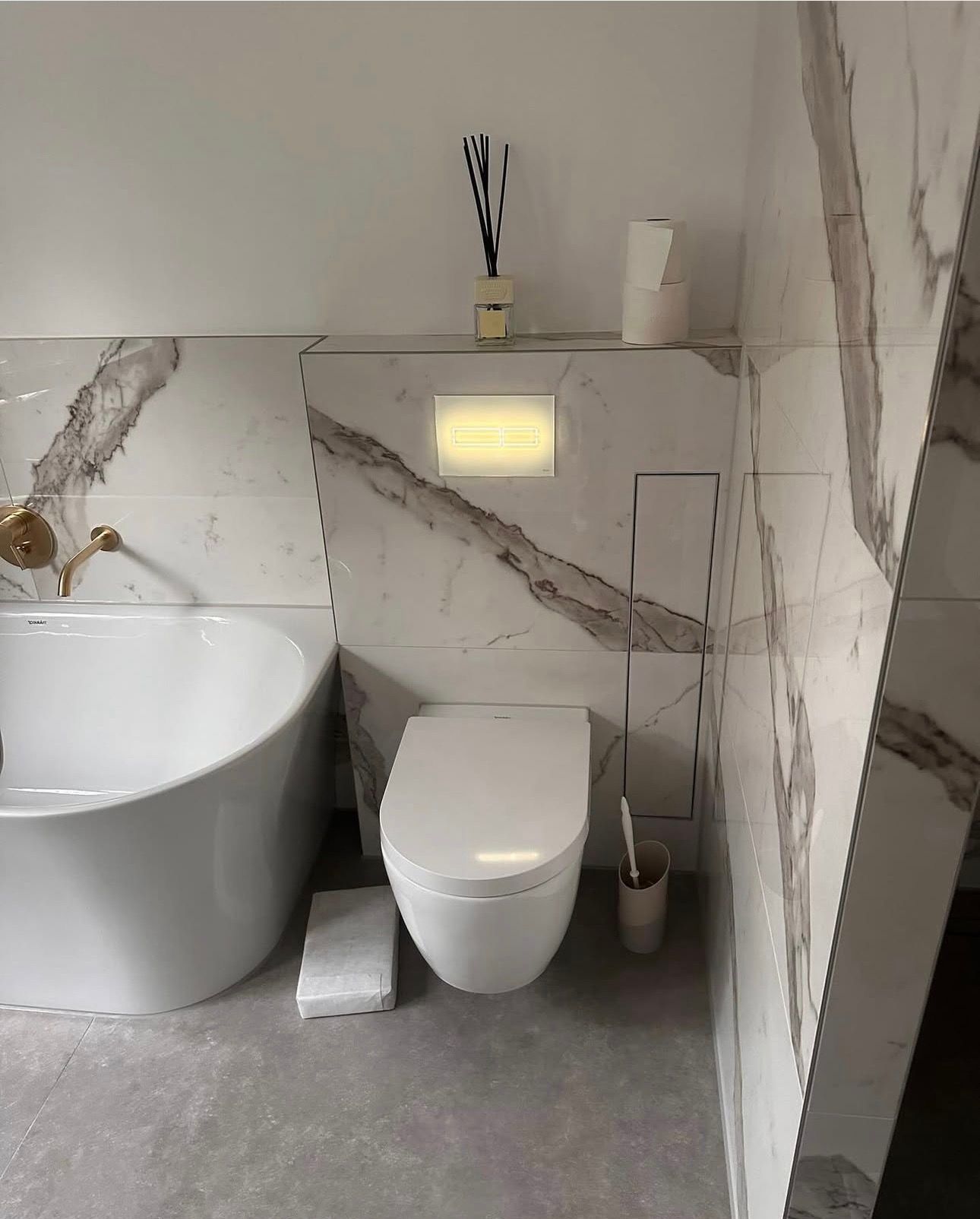 Weiße Toilette in einem mit Marmor gefliesten Badezimmer. Links befindet sich eine Badewanne.