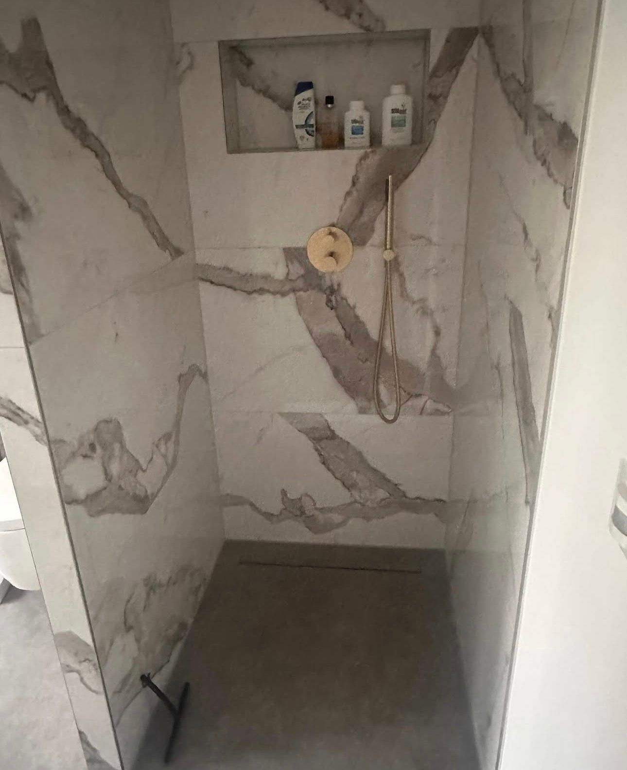 Dusche mit Wänden aus marmorgemusterten Fliesen, goldenem Duschkopf, eingebautem Regal mit Toilettenartikeln und Betonboden.