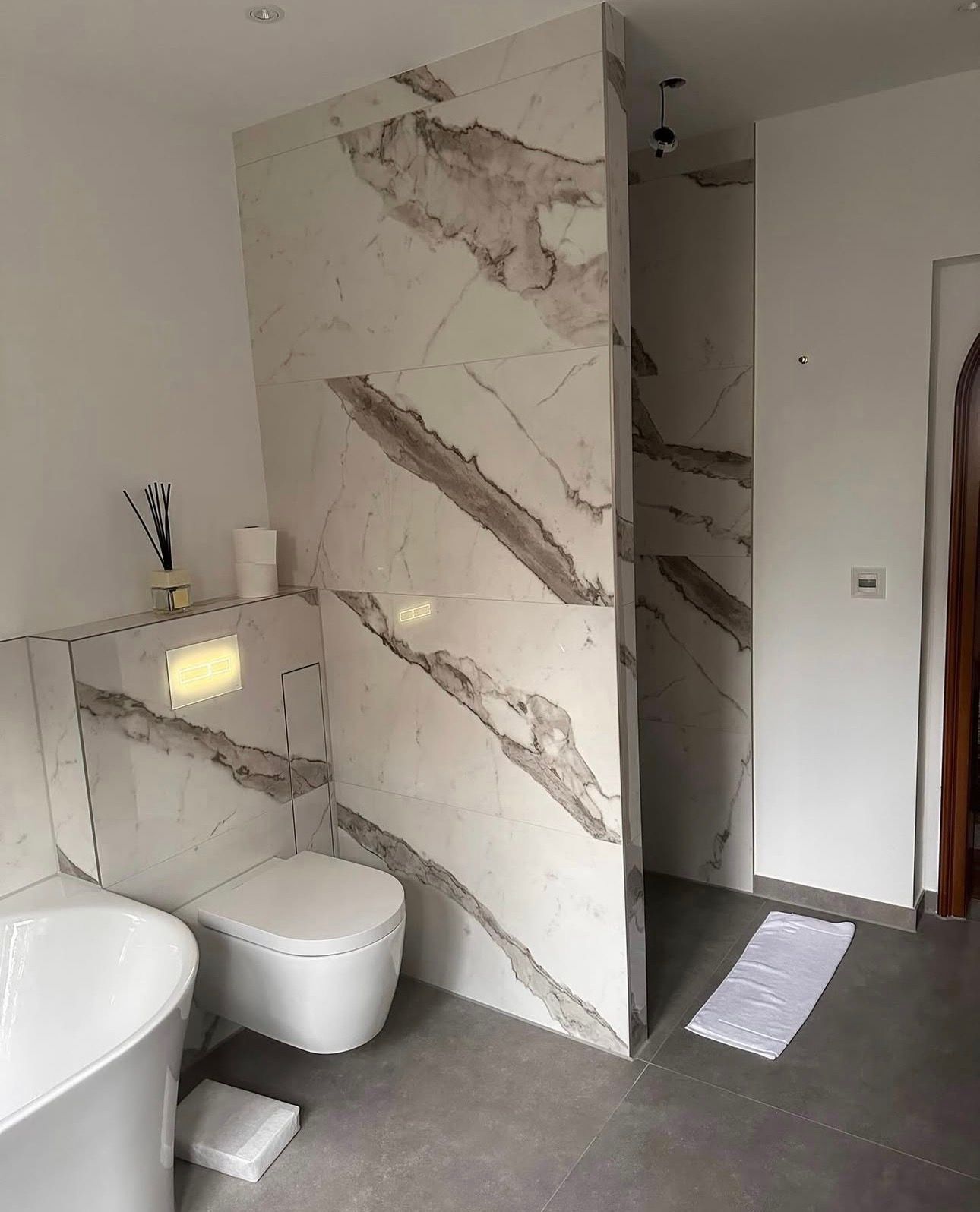 Modernes Badezimmer mit Wandfliesen in Marmoroptik, Toilette, Badewanne und Duschbereich; grauer Boden.