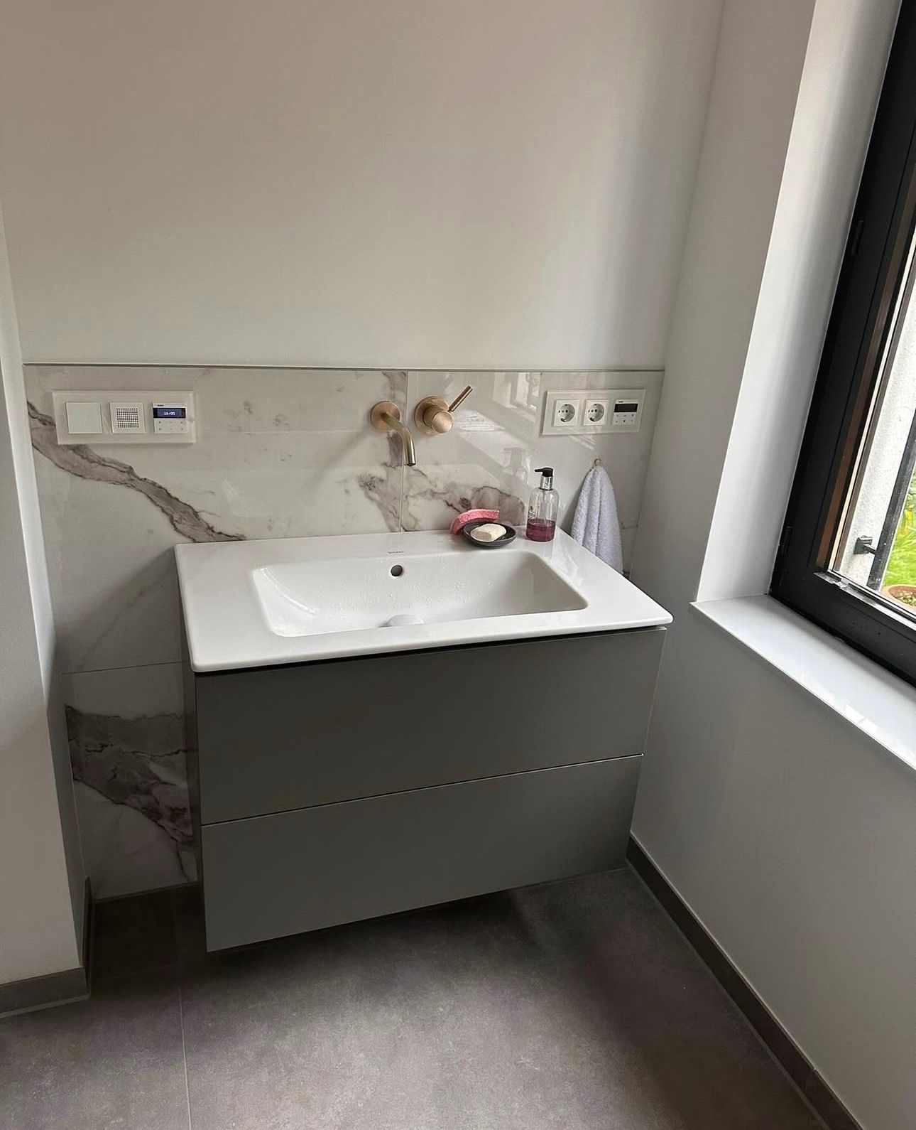 Badezimmer mit grauem Waschtisch, weißem Waschbecken, Rückwand in Marmoroptik und Fenster auf der rechten Seite.