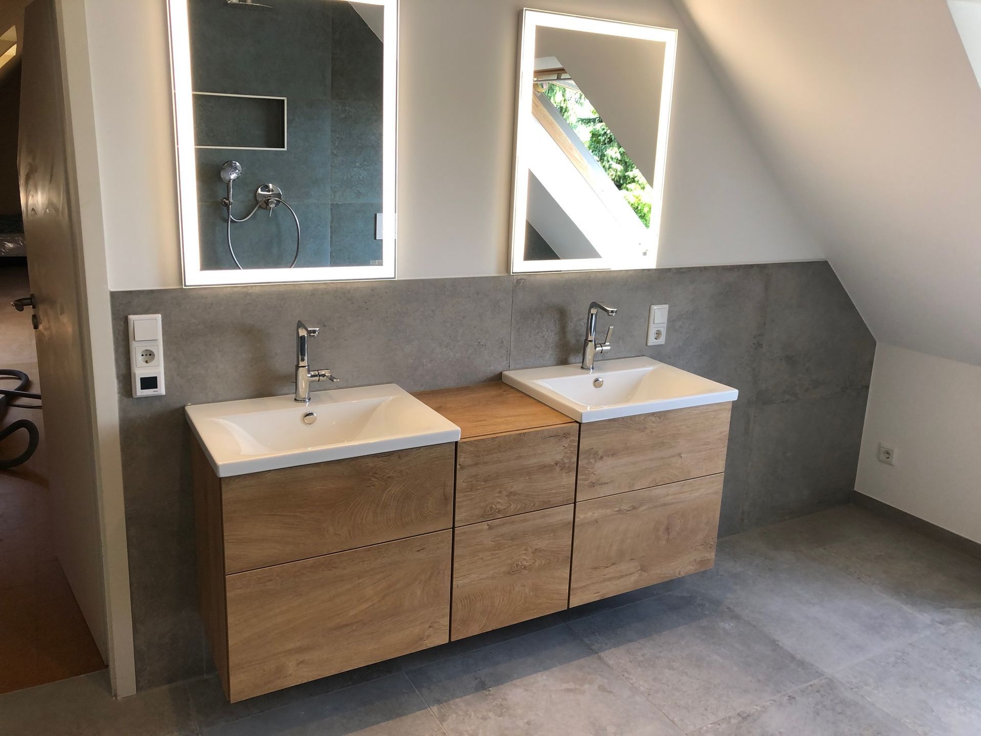 Badezimmer mit zwei Waschbecken, zwei beleuchteten Spiegeln, grauen und holzfarbenen Schränken sowie grauen Wänden und Boden.