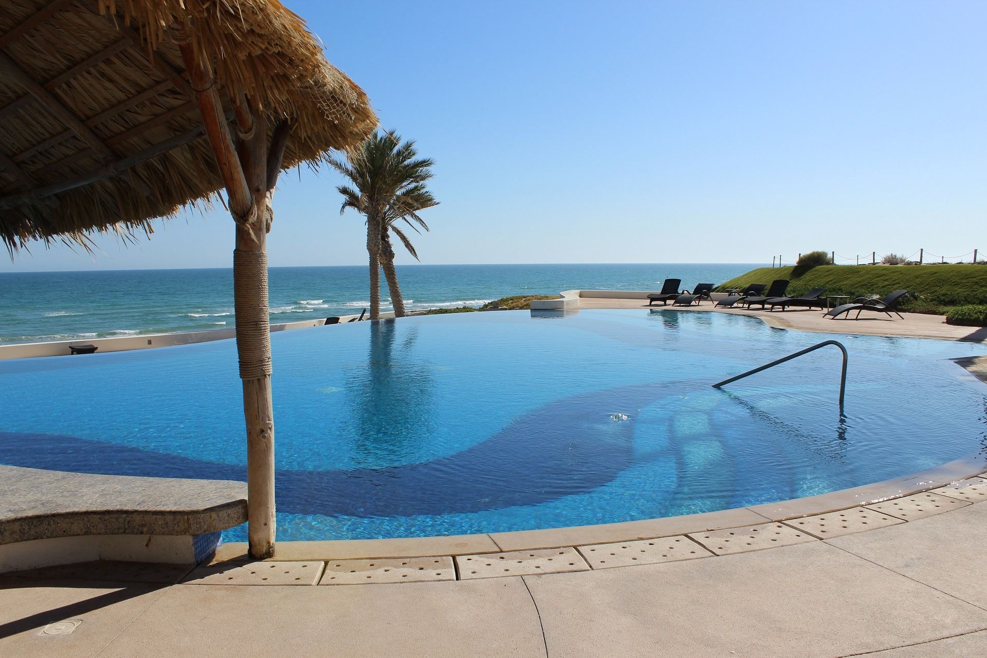 Oceanfront infinity pool beside the sea under a thatched shade, with lounge chairs and clear blue sky