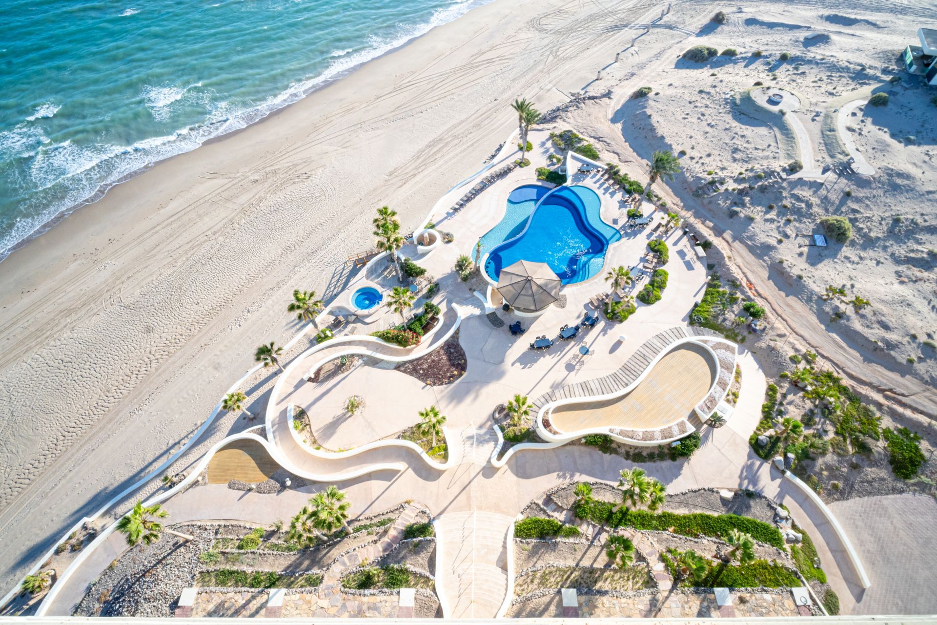Aerial view of a beachfront resort with a blue pool, winding paths, and sandy shoreline.
