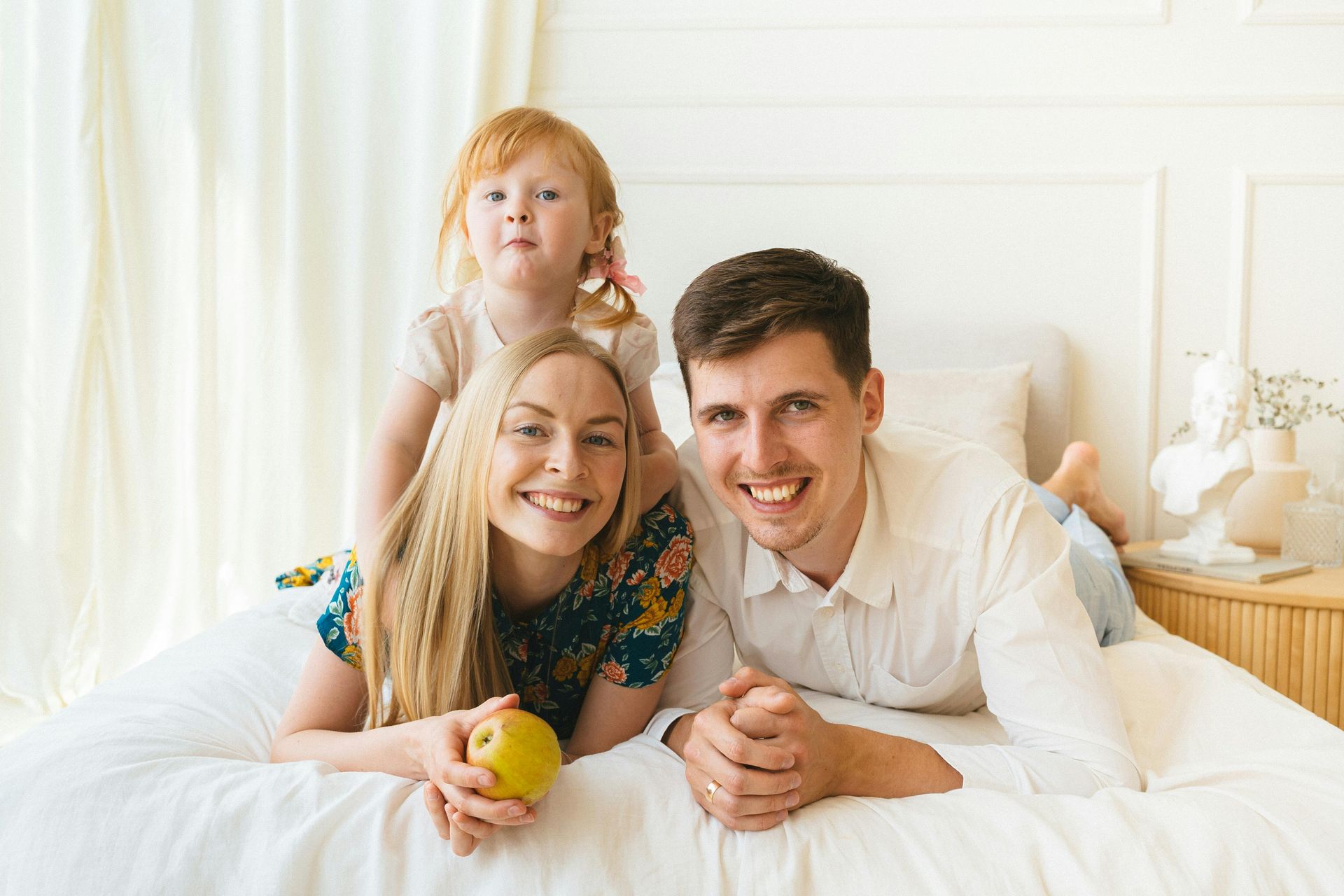 Family of three lying on a white bed, smiling at the camera with a child holding an apple