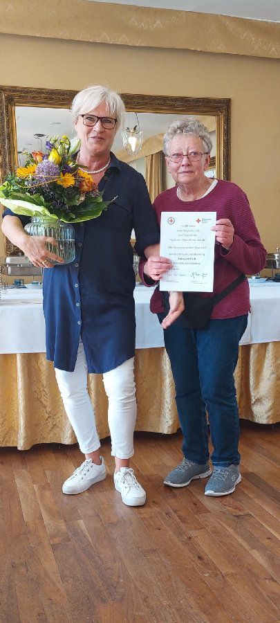Zwei Frauen stehen nebeneinander in einem Raum und halten Blumen und eine Urkunde in den Händen.