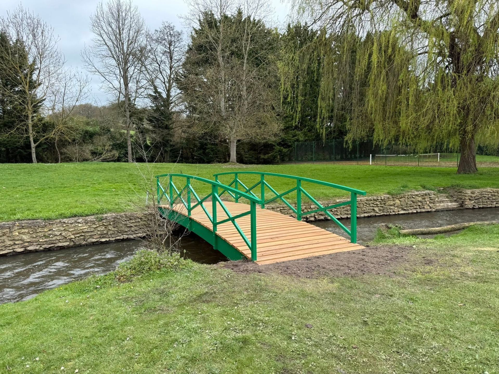 Passerelle piétonne verte et marron enjambant un petit ruisseau dans un parc herbeux.