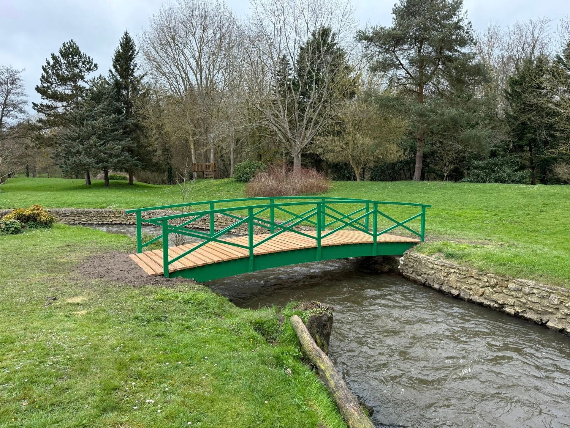 Passerelle piétonne verte enjambant un ruisseau dans un parc herbeux.