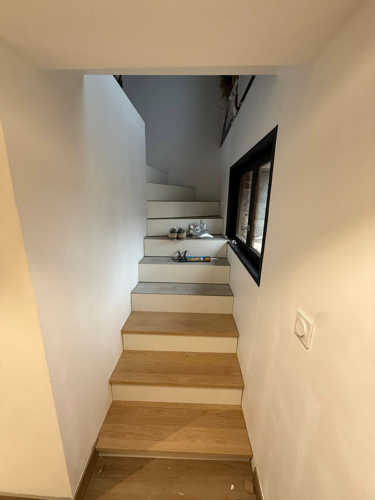Escalier aux marches claires et aux murs blancs ; une petite fenêtre se trouve sur le côté.
