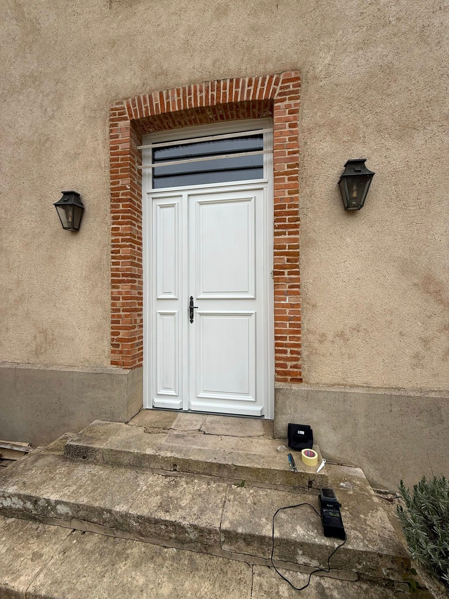 Porte blanche avec encadrement en briques, deux appliques sur un mur beige et marches en béton.