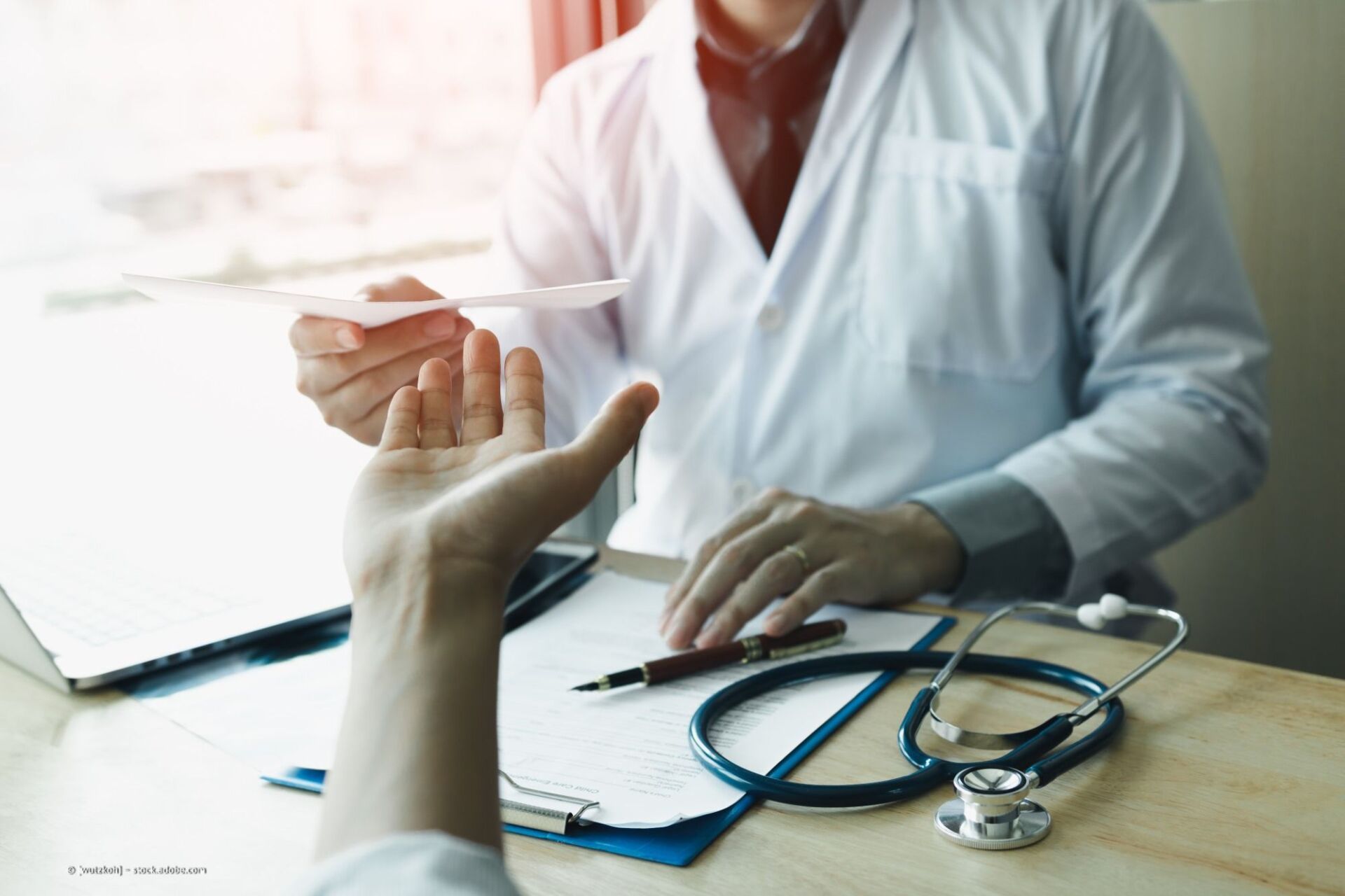 Arzt überreicht einem Patienten an einem Schreibtisch mit einem Stethoskop ein Rezept.