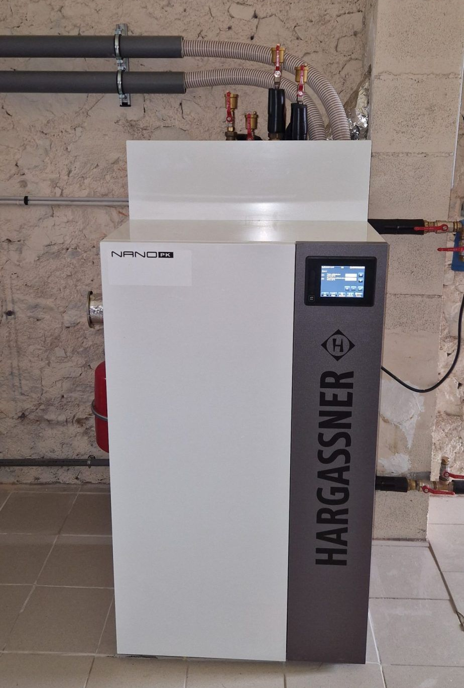 Une chaudière à granulés de bois Hargassner installée dans un sous-sol. Blanche et grise, avec un affichage numérique.