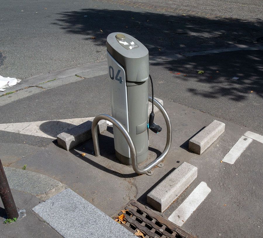 Borne de recharge pour véhicules électriques sur un trottoir, protégée par une barre métallique.