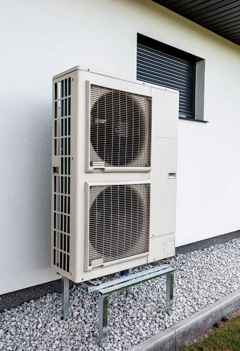 Unité de pompe à chaleur extérieure montée sur un support métallique, contre un bâtiment blanc avec une petite fenêtre.