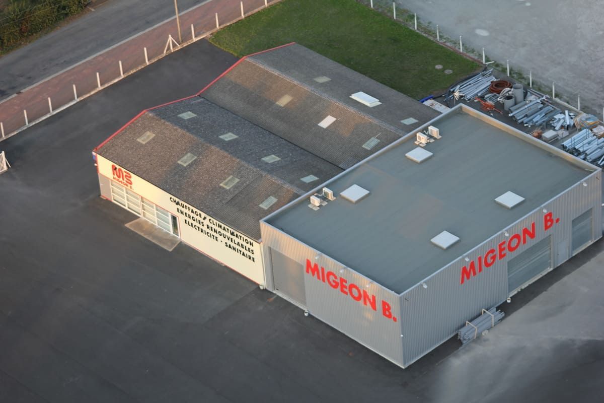 Vue aérienne de deux bâtiments industriels. L'enseigne « Migeon B. » orne le bâtiment à toit plat et façade argentée. Le bâtiment plus ancien possède un toit à pignon et une enseigne.