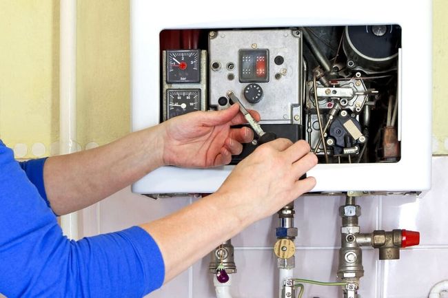 Une personne répare une chaudière à gaz ; ses mains tiennent un outil à l’intérieur de l’appareil ouvert. Tuyaux et manomètres visibles.