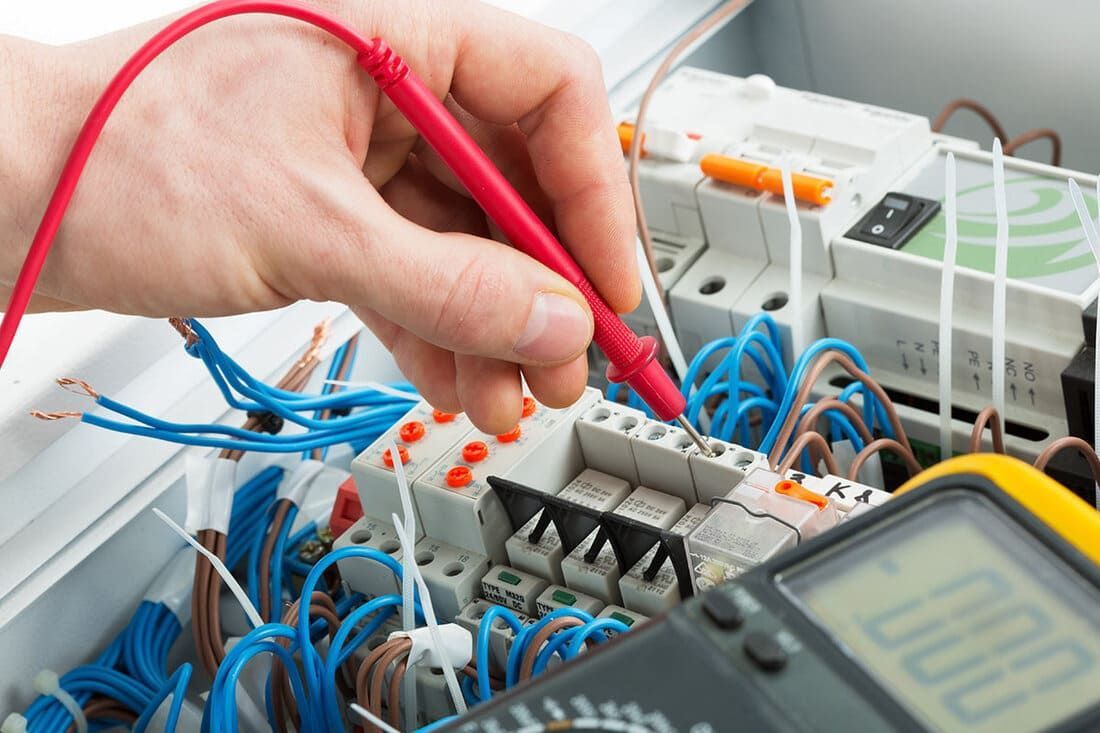 Une personne utilise une sonde rouge sur un tableau électrique pour tester le câblage avec un multimètre. Des fils bleus et blancs sont visibles.