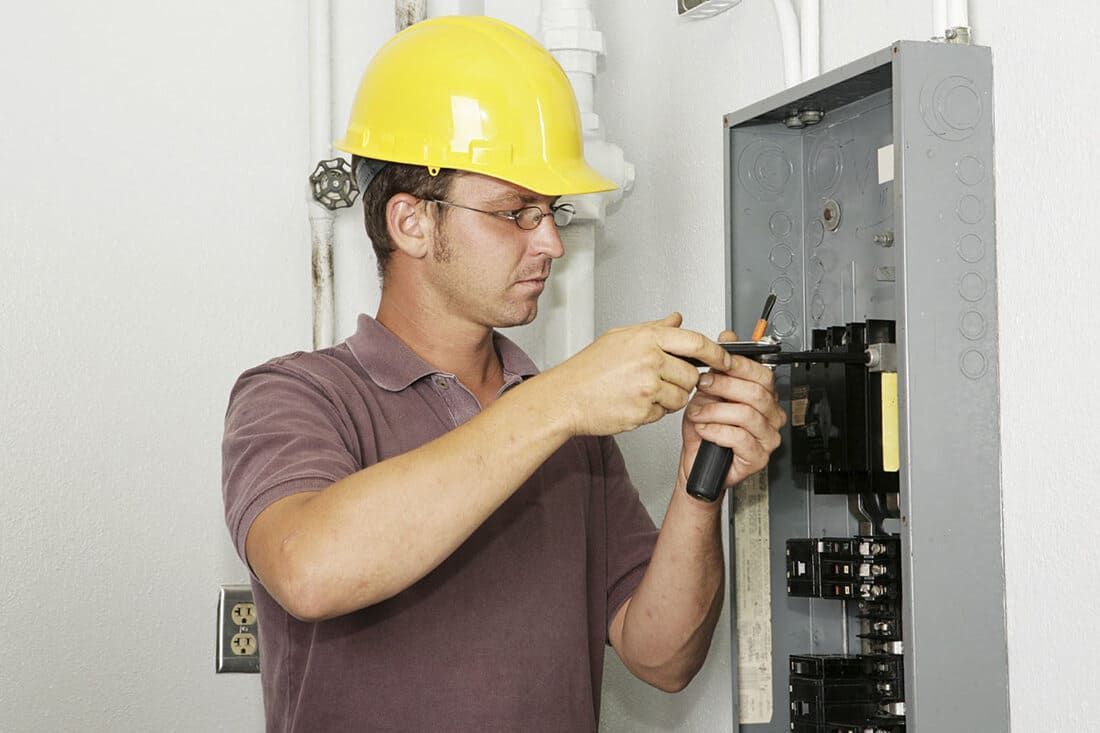 Électricien portant un casque de chantier jaune travaillant sur un tableau électrique avec un tournevis.