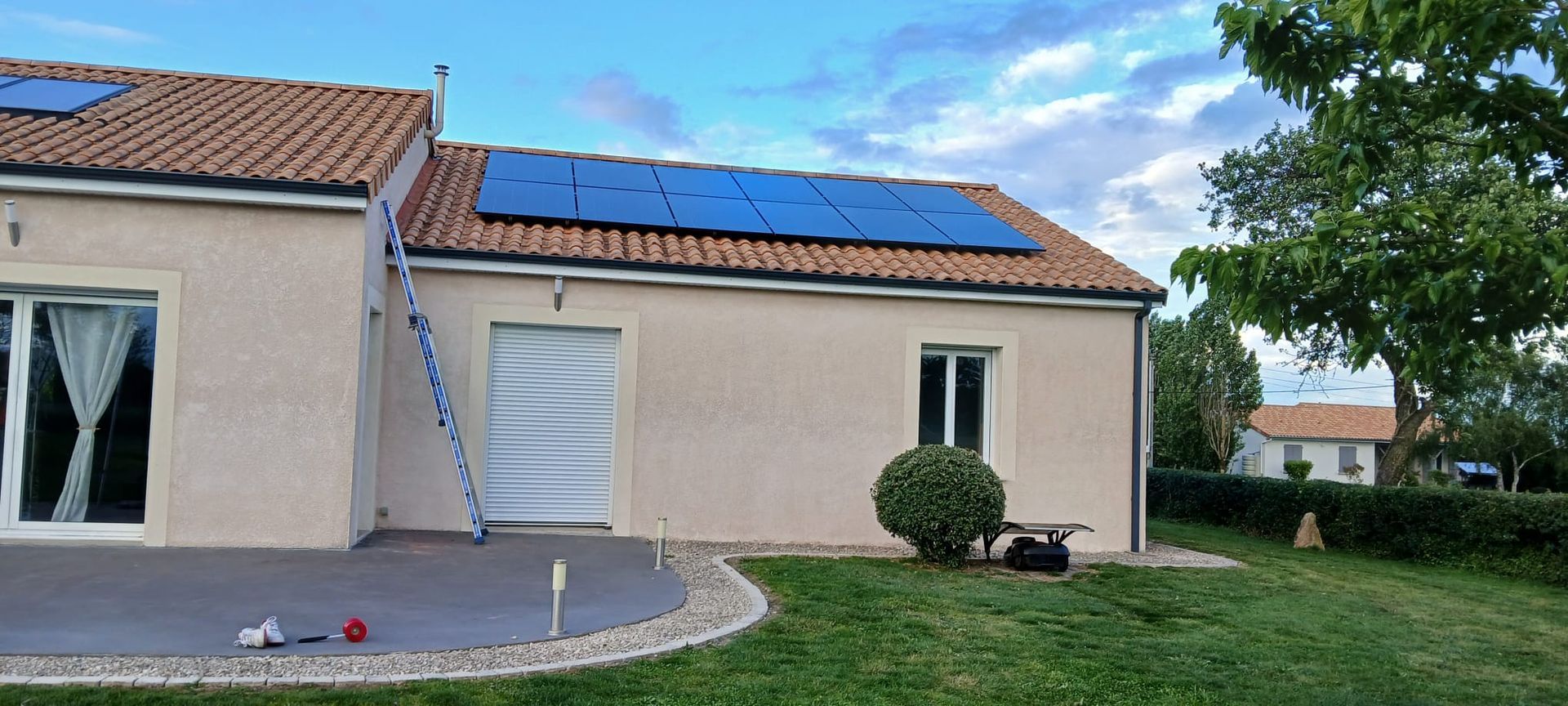 Maison beige de plain-pied avec toit de tuiles rouges, panneaux solaires et pelouse devant la maison sous un ciel bleu.