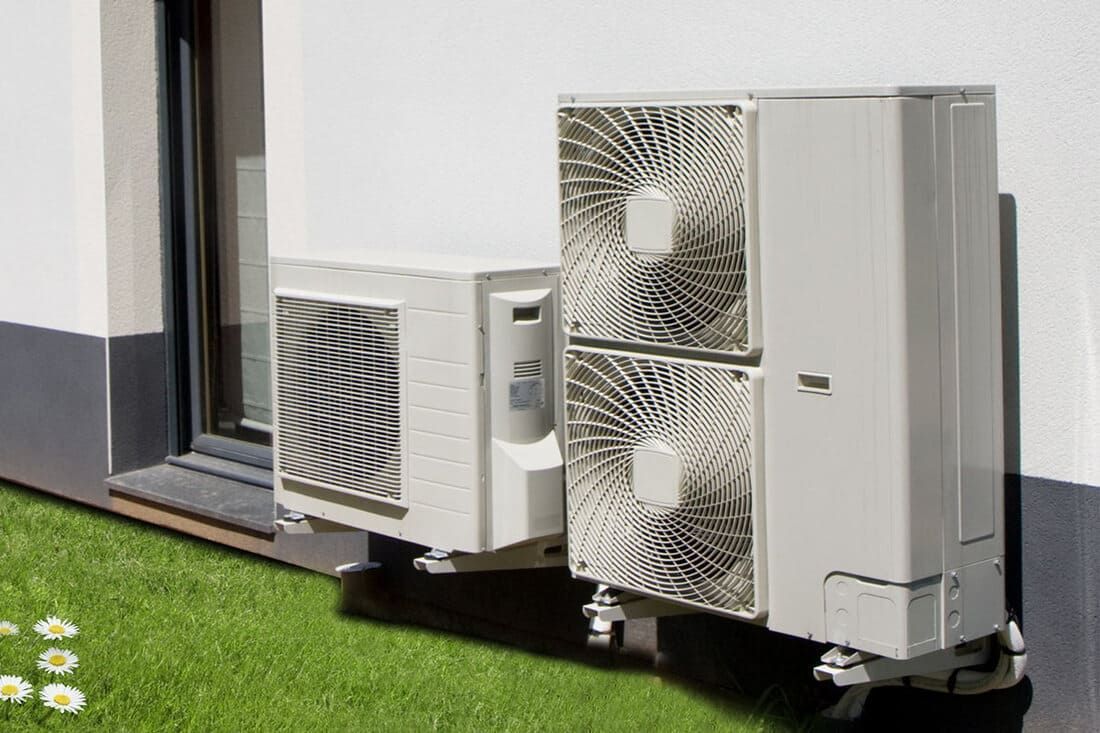 Deux unités de climatisation extérieures fixées à un mur blanc d'un bâtiment, au-dessus d'une parcelle de pelouse verte.