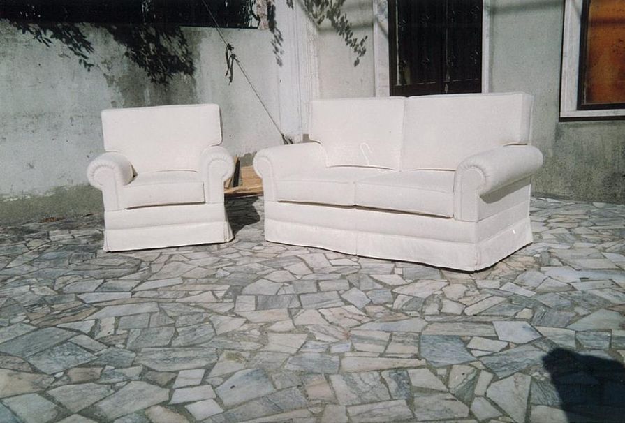 Uma poltrona branca e um sofá de dois lugares ficam em um pátio de pedra. O cenário é ao ar livre.