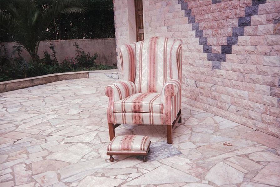 Poltrona com encosto alto e pufe com listras vermelhas e brancas em um pátio de pedra ao lado de uma parede texturizada.