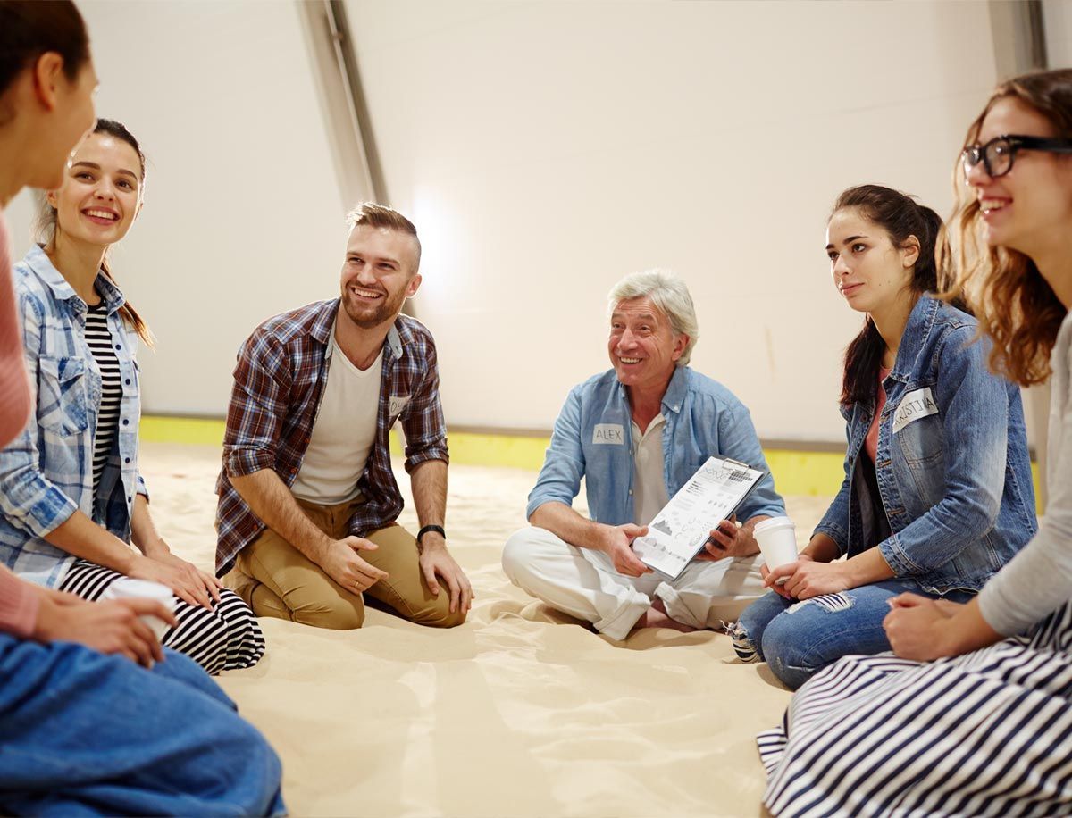 Un grupo de personas sentadas en círculo en el suelo, charlando y sonriendo en una habitación luminosa.