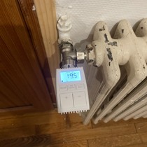 Thermostat numérique blanc sur un radiateur, affichant 19,5 °C. Installé près d'un cadre de porte en bois et d'un mur blanc.