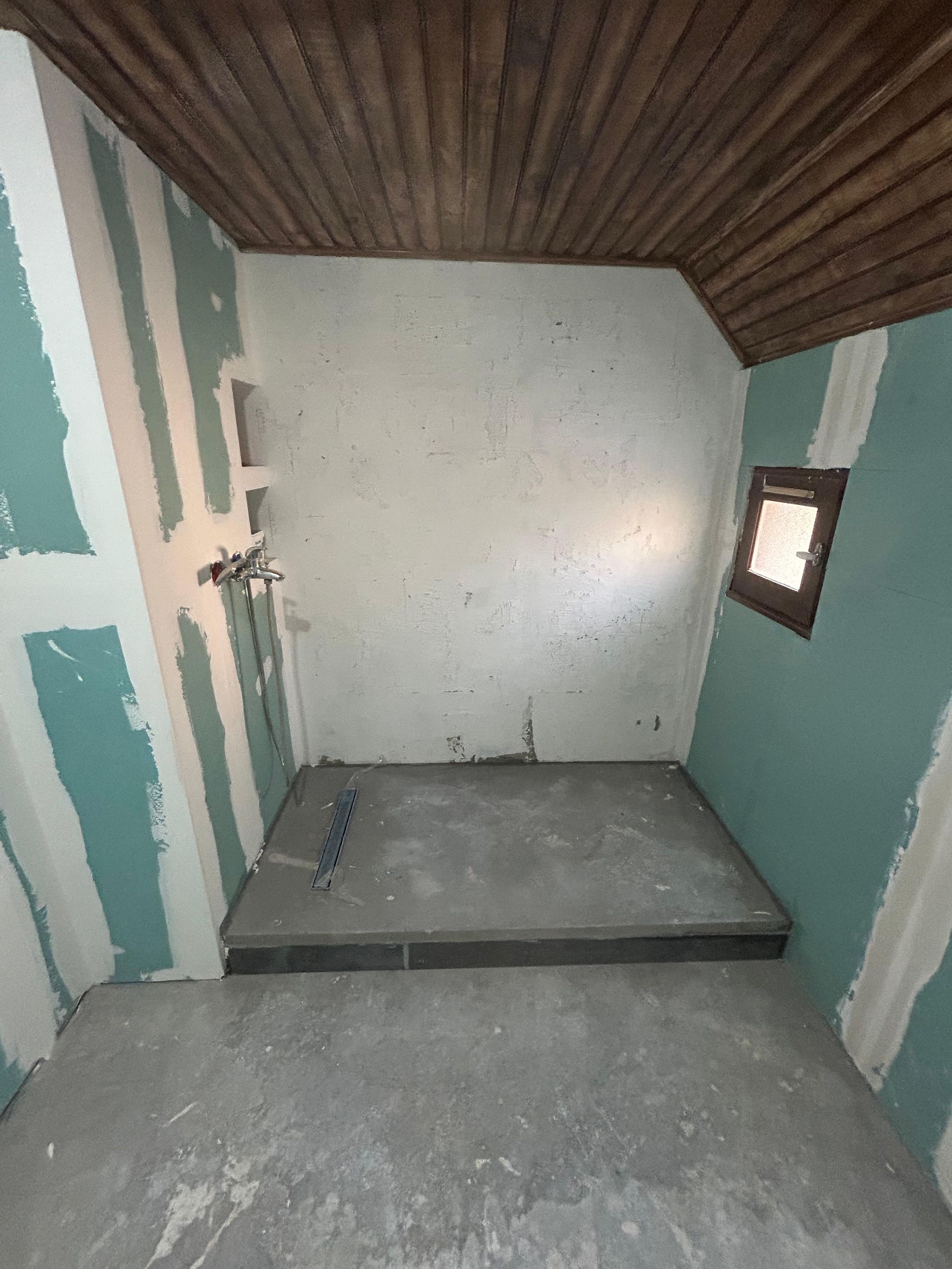Petite salle de bain en rénovation, sol de douche gris, murs verts, plafond en bois apparent, petite fenêtre.